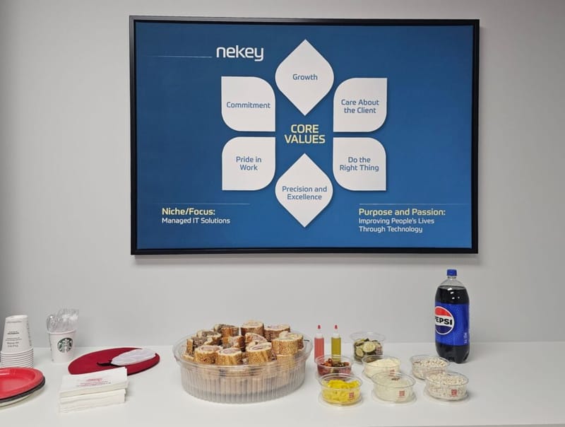 Nekey core values slide on a wall-mounted screen above a table with a sandwich platter, condiments, and a Pepsi bottle at a company event