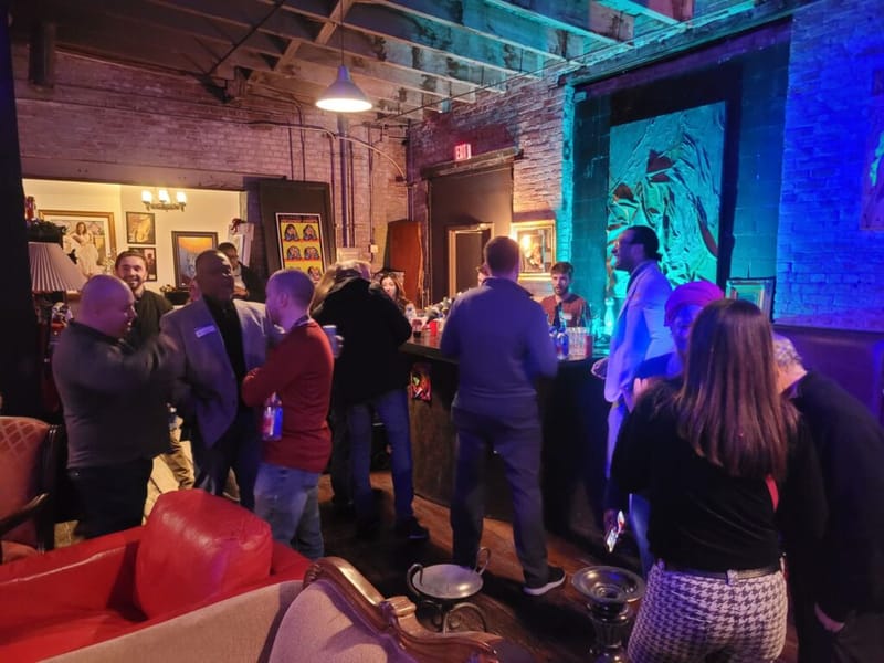 Attendees mingling near a bar at a dimly lit venue with exposed brick walls and colorful stage lighting during a networking event