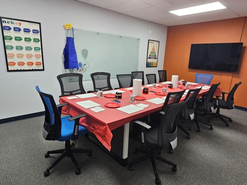 conference room set up for a paint and sip event, long table with red tablecloth, paint supplies, mugs, and paper at each seat, blue apron hanging on whiteboard, nekey posters on walls